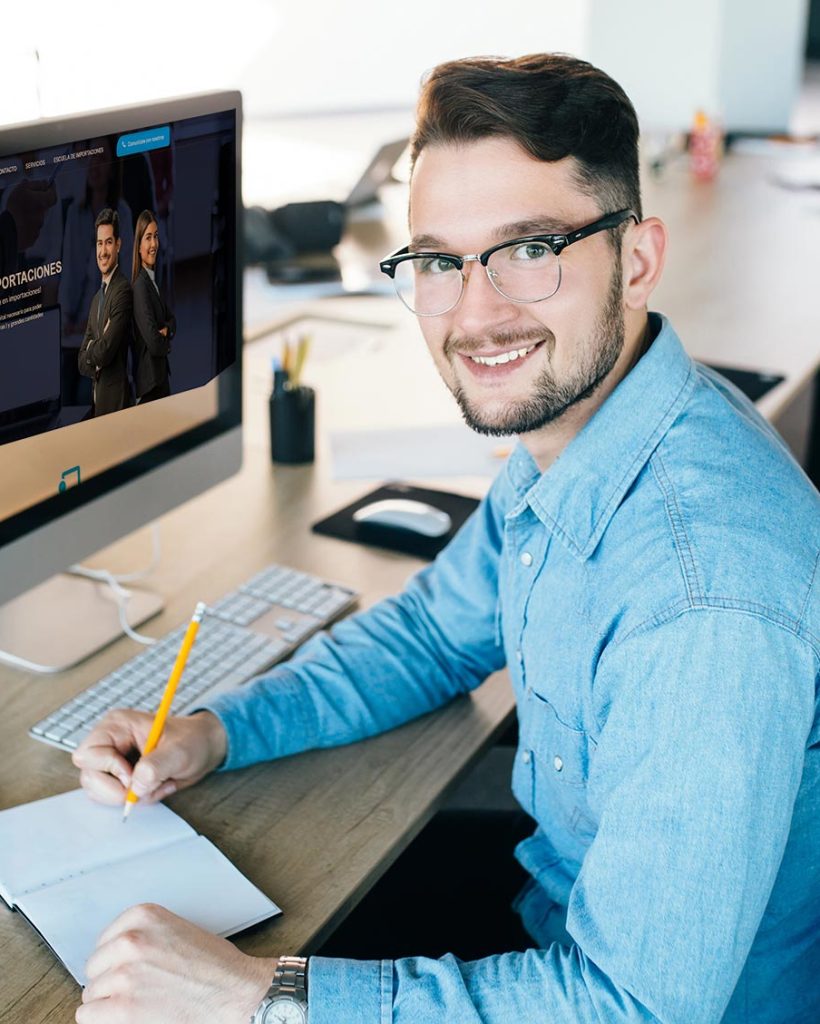 hombre-joven-glassess-trabajando-su-lugar-trabajo-oficina-viste-camisa-azul-escribiendo-cuaderno-sonriendo-camara